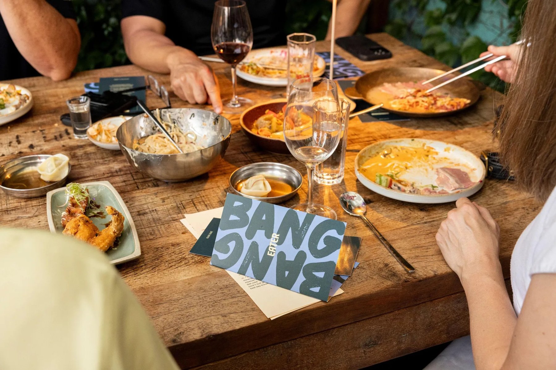 Hands around a shared table at Eater Bang Bang, with noodles, dumplings, and a Bang Bang card