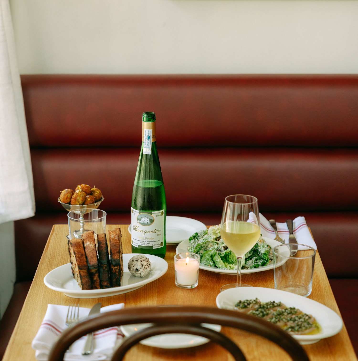 A set table at Marée: txakoli, grilled bread, salad, and crudo against a red booth